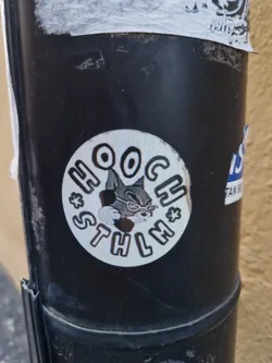 A round black and white sticker featuring a cartoon cat wearing sunglasses. The text 'HOOCH * STHLM *' surrounds the cat. The sticker is affixed to a dark-colored cylindrical object, possibly a drainpipe, outdoors. The style is reminiscent of vintage cartoons or graphic design.