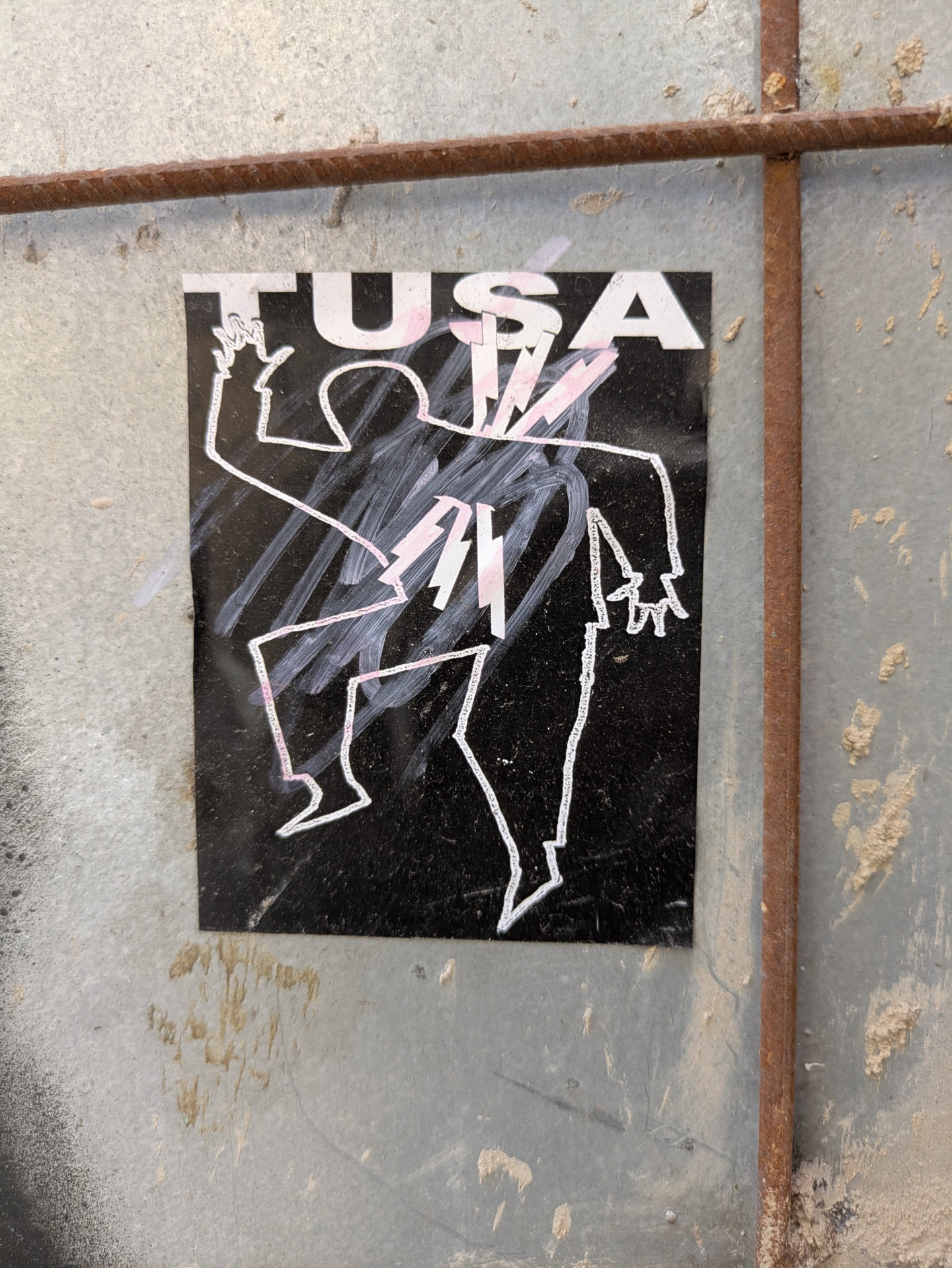 This black sticker features a white outline of a dancing or gesturing figure. The figure is stylized and abstract, with jagged lines and sharp angles. Above the figure, the word TUSA is printed in bold, white letters. The sticker is affixed to a weathered metal surface.