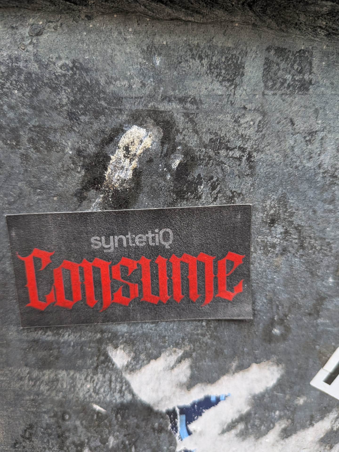 A rectangular sticker with a dark grey, slightly textured background. The prominent word 'CONSUME' is displayed in a vibrant red, gothic-style font, dominating the lower half of the sticker. Above it, in smaller white sans-serif text, is the word 'syntetiQ'. The sticker shows minor signs of wear, with slight scuffs and a clear, crisp edge, indicating it's a physical adhesive item. It is affixed to a heavily textured, dark, possibly metallic or concrete wall, which is mottled with lighter patches and grime.