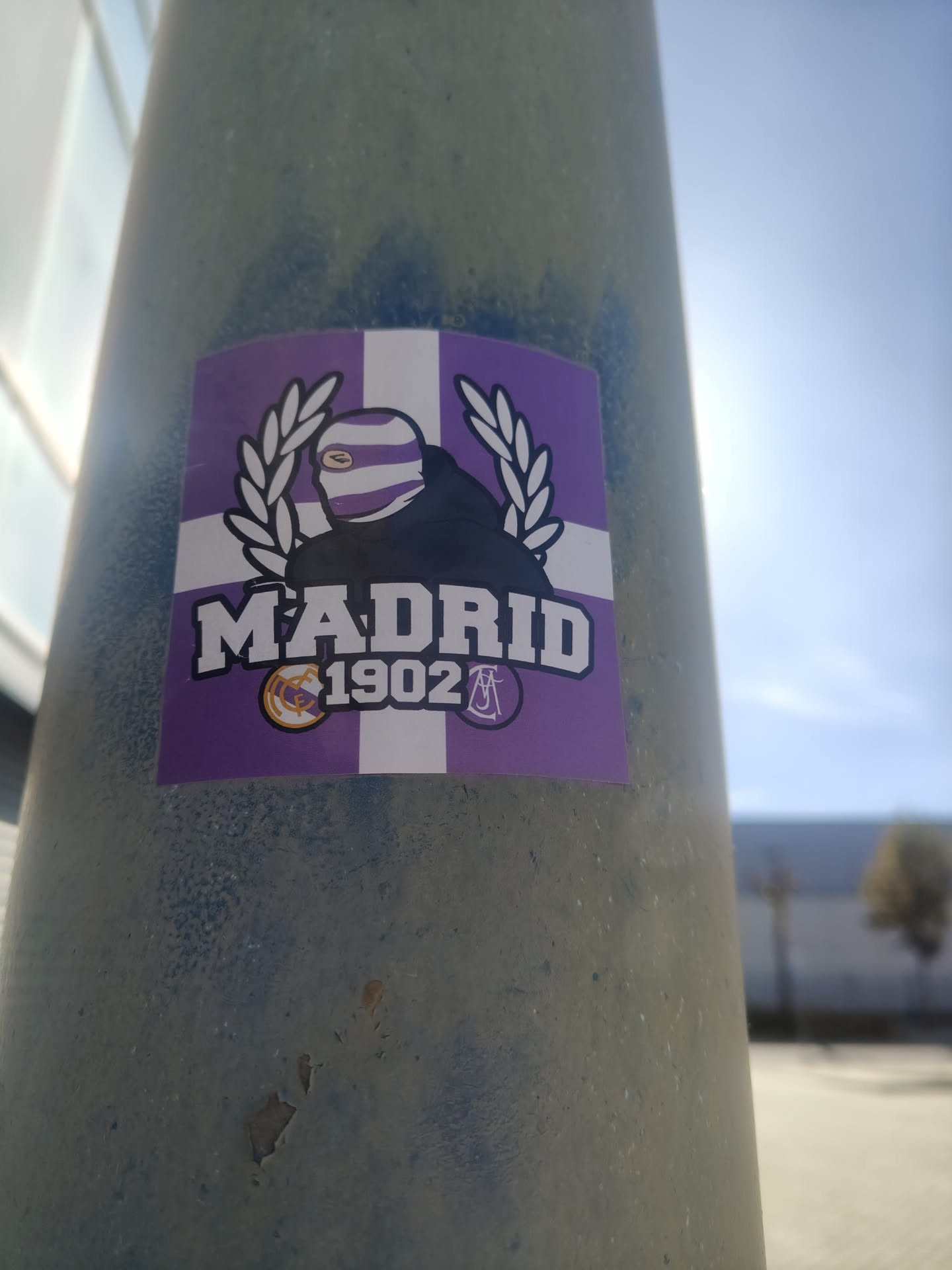 A rectangular sticker featuring a central design on a background of alternating purple and white quadrants. The main element is a stylized figure wearing a black hooded garment and a purple and white striped mask, with only one eye visible. The figure is flanked by white laurel wreaths. Below the figure, in bold white letters with a black outline, is the word 'MADRID', and beneath it, '1902'. To the left of '1902' is a small, circular Real Madrid crest in gold and purple. To the right of '1902' is another small, circular logo on a purple background, featuring stylized white letters resembling 'US', commonly associated with the Ultras Sur fan group.