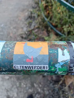 A sticker featuring a minimalist illustration of a goose, with the text "ENTENWERDER1" underneath. The sticker is affixed to a rusty green metal surface.