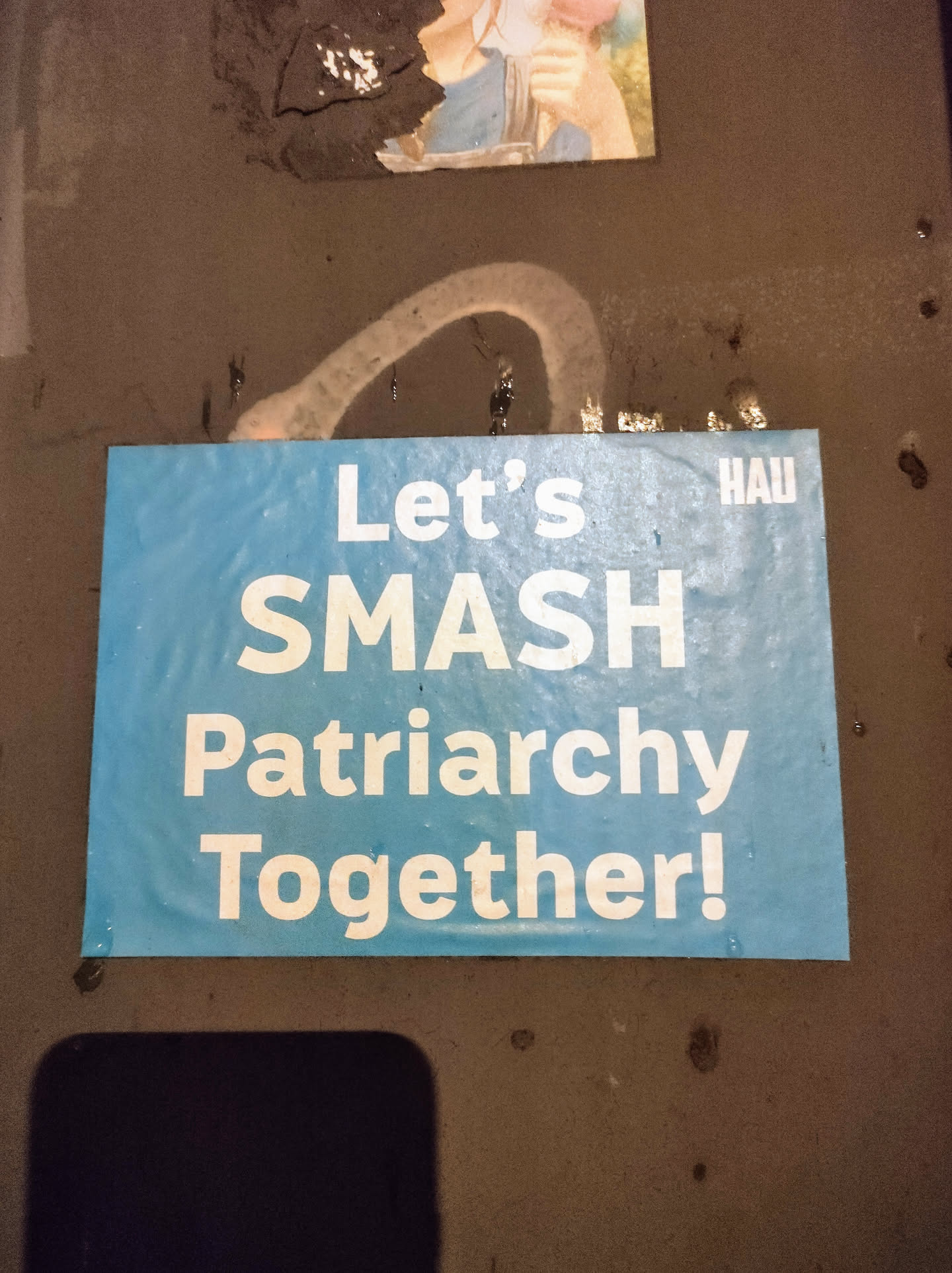 A light blue square sticker with the text "Let's Smash Patriarchy Together!" in white, bold letters. There is a small, barely visible logo in the top right corner.