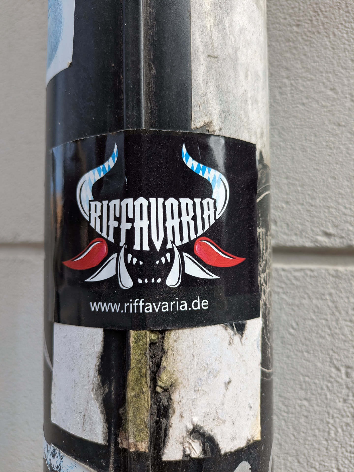 This sticker features the stylized text 'RIFFAVARIA' above a monster-like face with horns that resemble the Bavarian flag pattern. The URL www.riffavaria.de is printed below the design. The sticker is affixed to a pole.