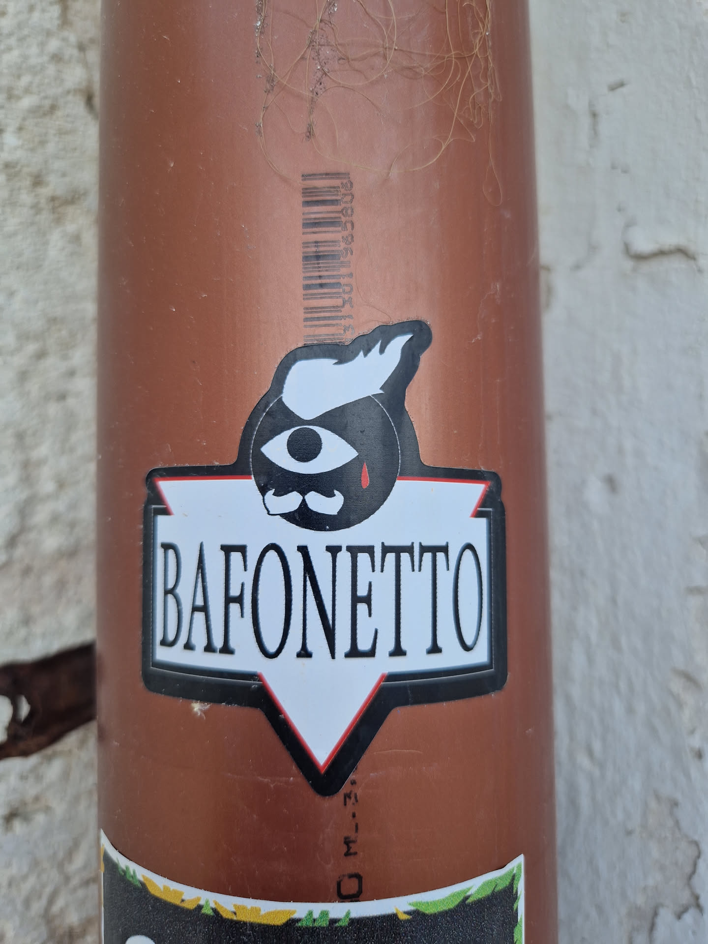 A black and white sticker with a stylized illustration of an eye with a mustache and a single tear. Below the illustration is the text "BAFONETTO" in a bold, sans-serif font.  The sticker is applied to a brown pipe.