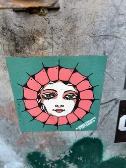A square sticker on a weathered surface features a drawing of a woman's face. The face has fair skin, blue eyes, and a small, red mouth. The face is framed by pink petals with black markings on the edges, resembling a flower.  The sticker has a green background. At the bottom right, the name "ALESSIO ERRANTE * TATTOOER *" is printed.