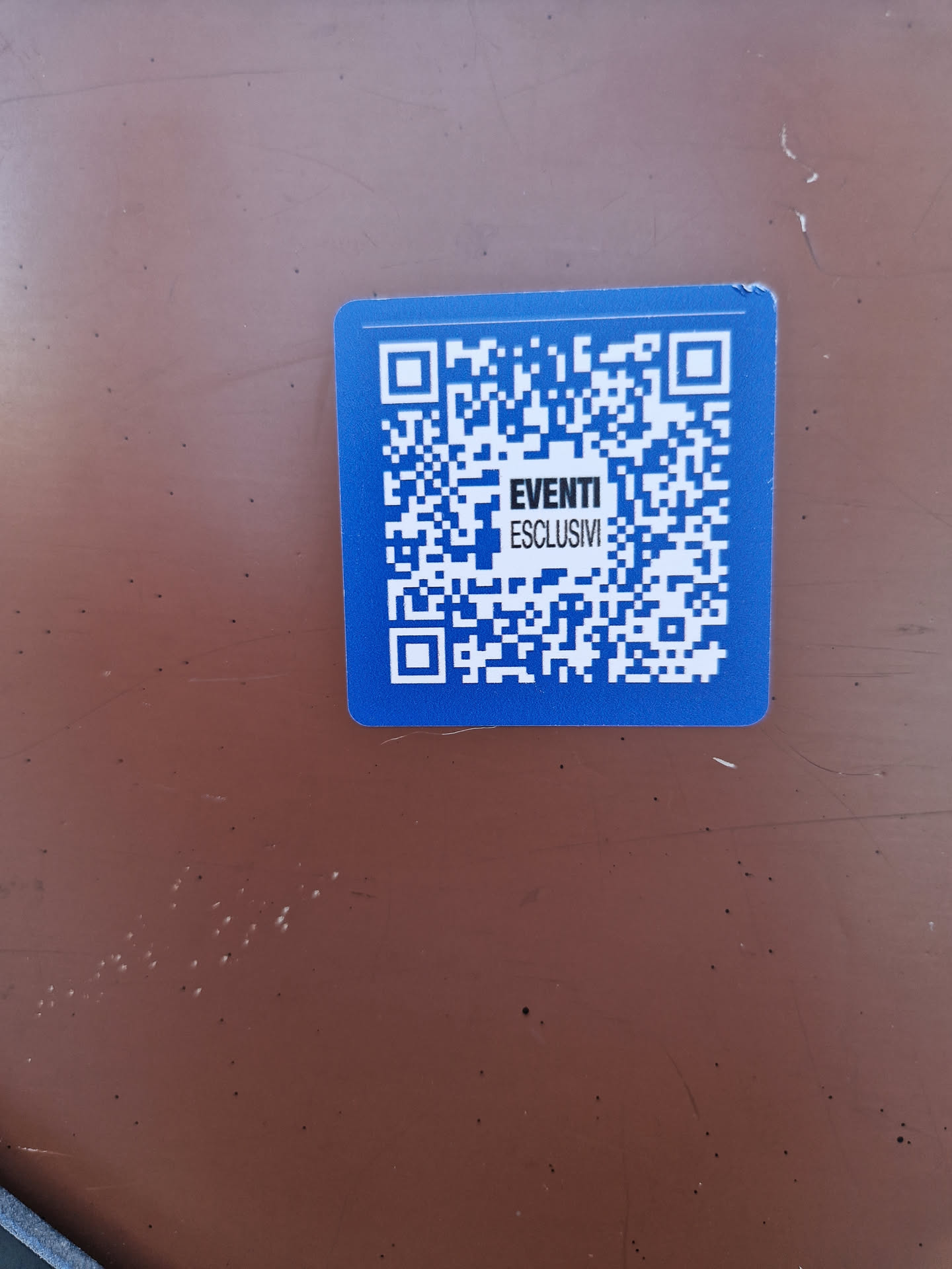 A blue square sticker with a QR code and the text "EVENTI ESCLUSIVI" (meaning "Exclusive Events" in Italian) in the center.  The sticker is affixed to a brown surface.