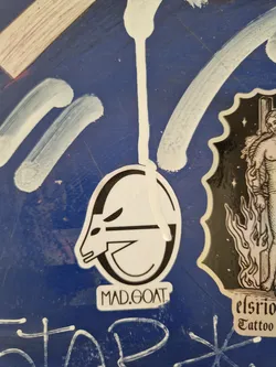 A sticker with a minimalist black and white illustration of a goat's head inside a circle. Underneath is the text 'MAD.GOAT'.
