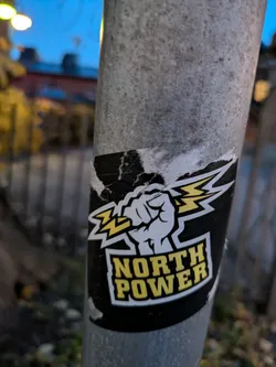 A worn sticker with a fist holding a lightning bolt and the words "NORTH POWER" in yellow letters on a black background. The sticker is affixed to a gray metal pole outdoors.