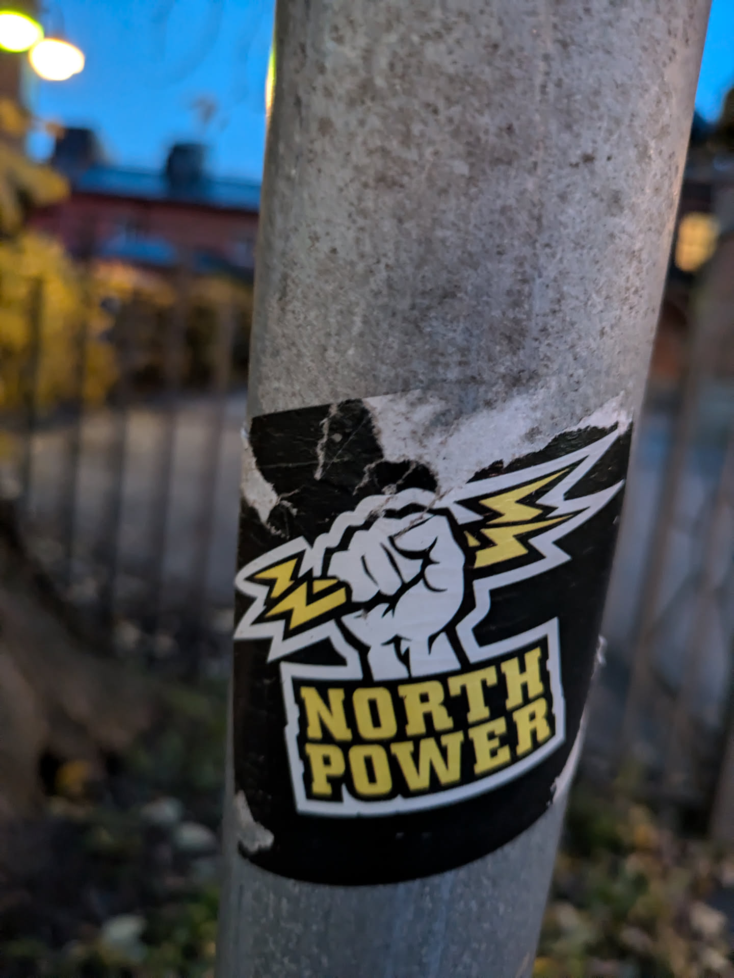 A worn sticker with a fist holding a lightning bolt and the words "NORTH POWER" in yellow letters on a black background. The sticker is affixed to a gray metal pole outdoors.