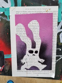 A purple and pink stencil graffiti of a face on a page of a book. The stencil is layered with different shades of purple and pink. The background is a page of a book with some text visible. The stencil is a simple design of a face with a nose, mouth and eyes.