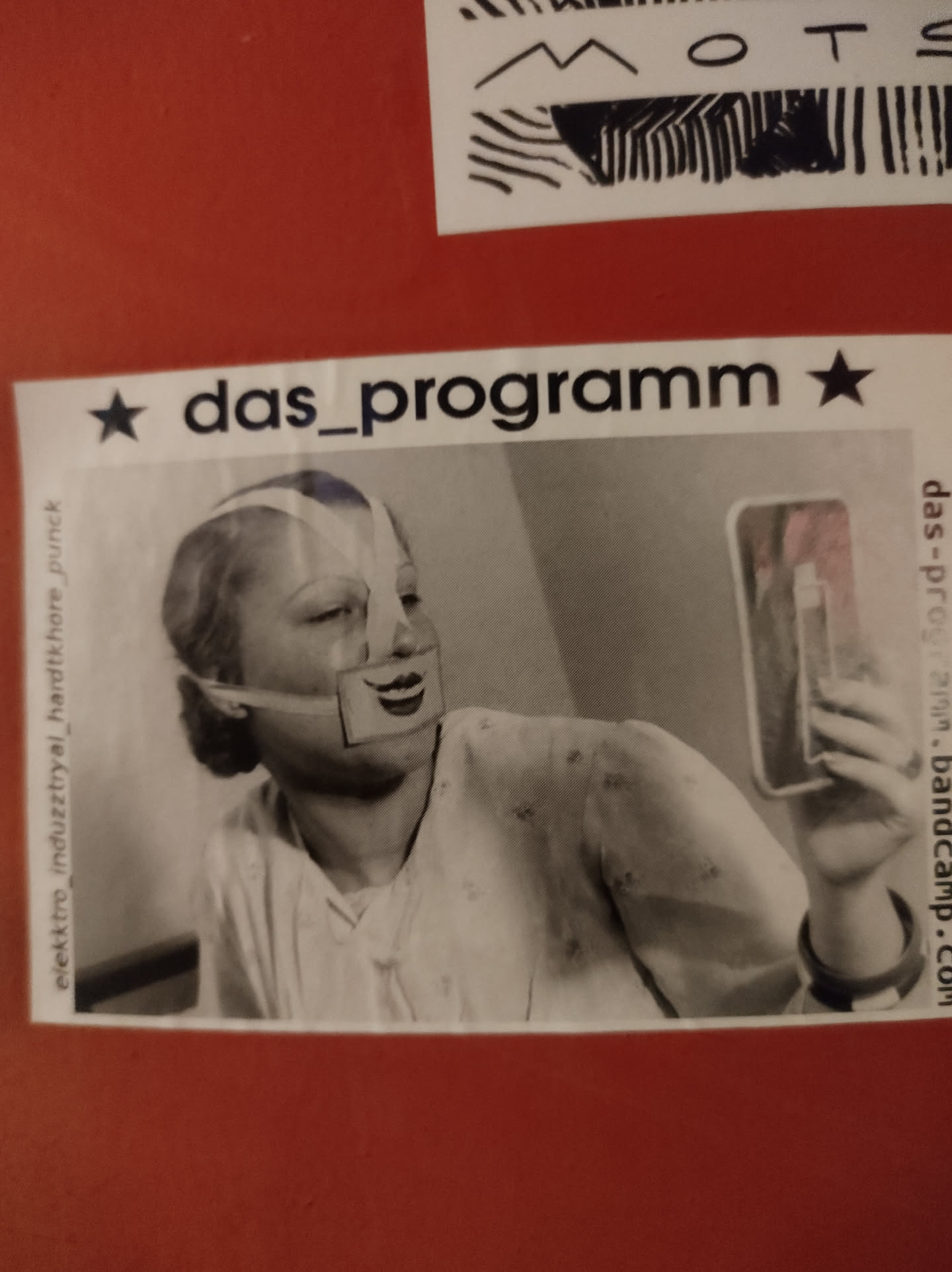 Black and white photo of a woman looking at herself in a mirror, wearing a mask. The sticker features the band's name and Bandcamp link. 