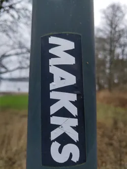 A close-up shot of a black and white MAKKS sticker affixed to a dark teal pole outdoors. The sticker features the word "MAKKS" in a stylized, slightly distorted sans-serif font. The background is blurred, showing some vegetation and trees.
