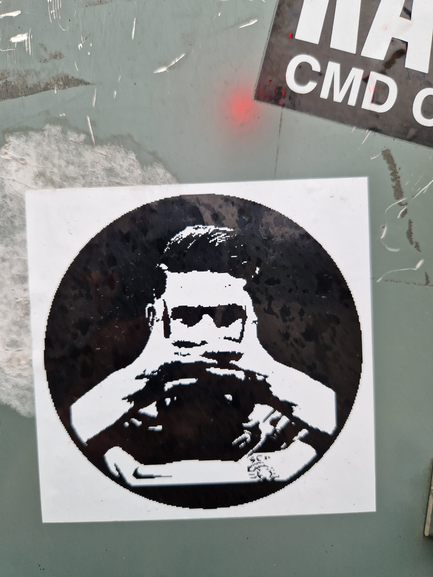 A monochrome round sticker featuring a stylized illustration of a person wearing sunglasses and appearing to be holding something in front of them. The style is reminiscent of stencil art or a screen print.  The image is predominantly black and white, with strong contrasts.