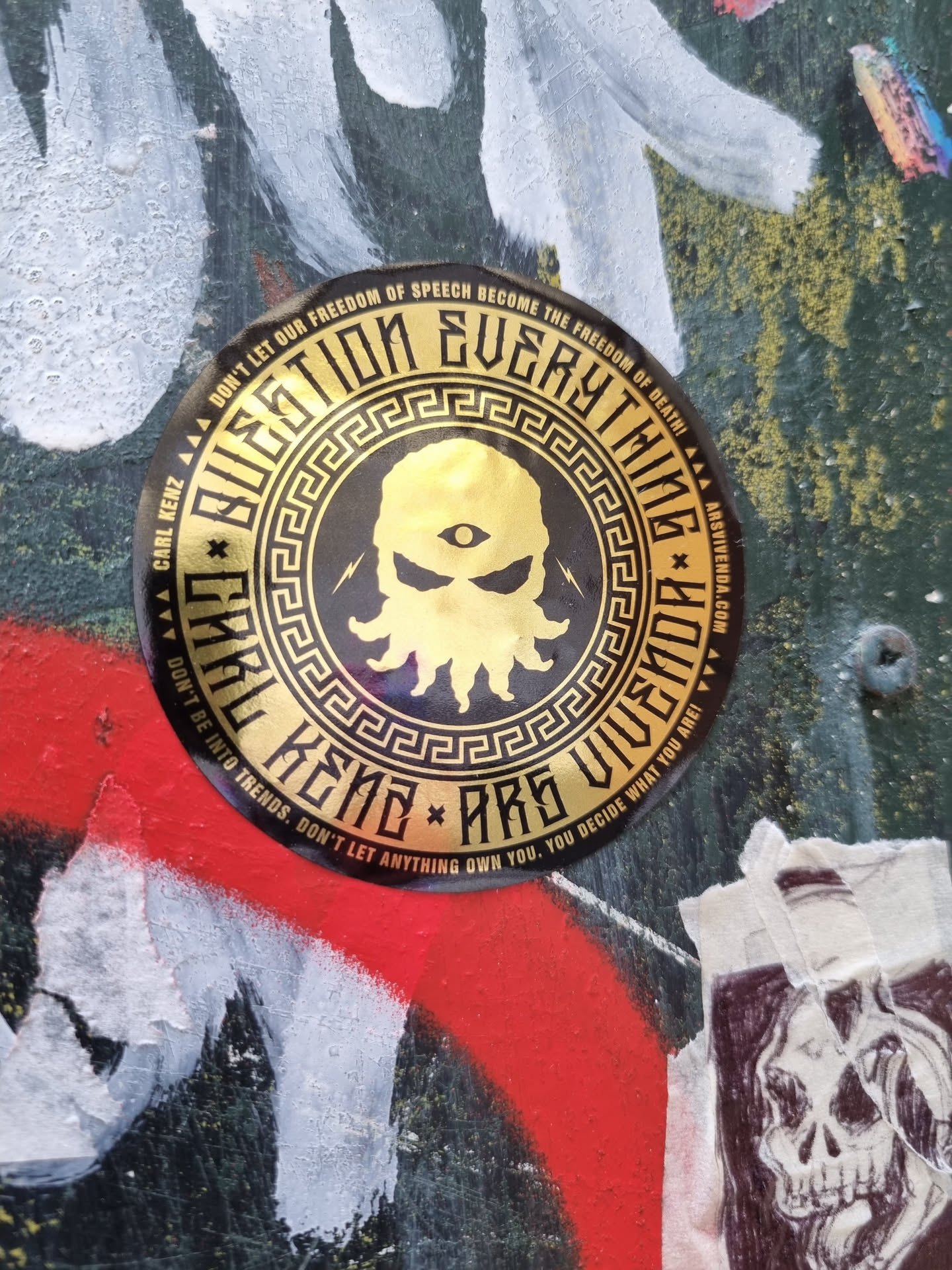 Gold and black round sticker with an octopus-like creature with one eye in the center, surrounded by a Greek key pattern and text. The text on the sticker promotes critical thinking and individuality.