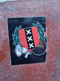 A black and red sticker depicting the coat of arms of Amsterdam, accompanied by a megaphone and a football.  The design is bold and graphic.