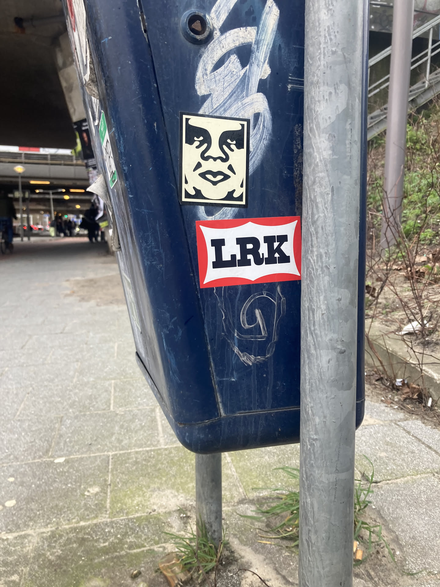 The sticker at the center of the image is a well-known 'Obey Giant' design, featuring a stylized black and white face with large eyes and an open mouth. It is placed on a dark blue, possibly metallic, surface. Below the Obey Giant sticker is another sticker with a red border and a white background, displaying the bold black letters 'LRK'. The overall presentation suggests urban art or graffiti culture.