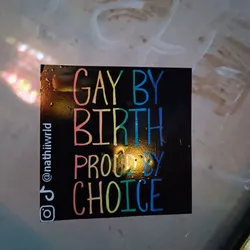 A black square sticker with rainbow-colored text. The text says "Gay by birth, proud by choice". There are also social media handles on the bottom left corner.
