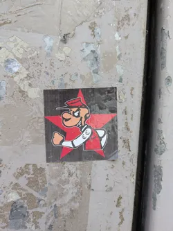 The sticker features a cartoon character with an orange face and a sinister expression, wearing a red cap and uniform with white trim. The character is posed against a bright red star on a dark grey background. The sticker is affixed to a peeling painted surface.