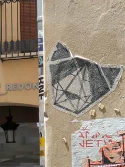A geometric sticker adhered to a beige, textured wall. The sticker is grayscale and features a pattern of connected lines forming abstract shapes, resembling facets of a gem. Some small, rectangular pieces of paper or residue are scattered around the sticker. Below it, a partially torn poster with red text and graphic elements is visible.