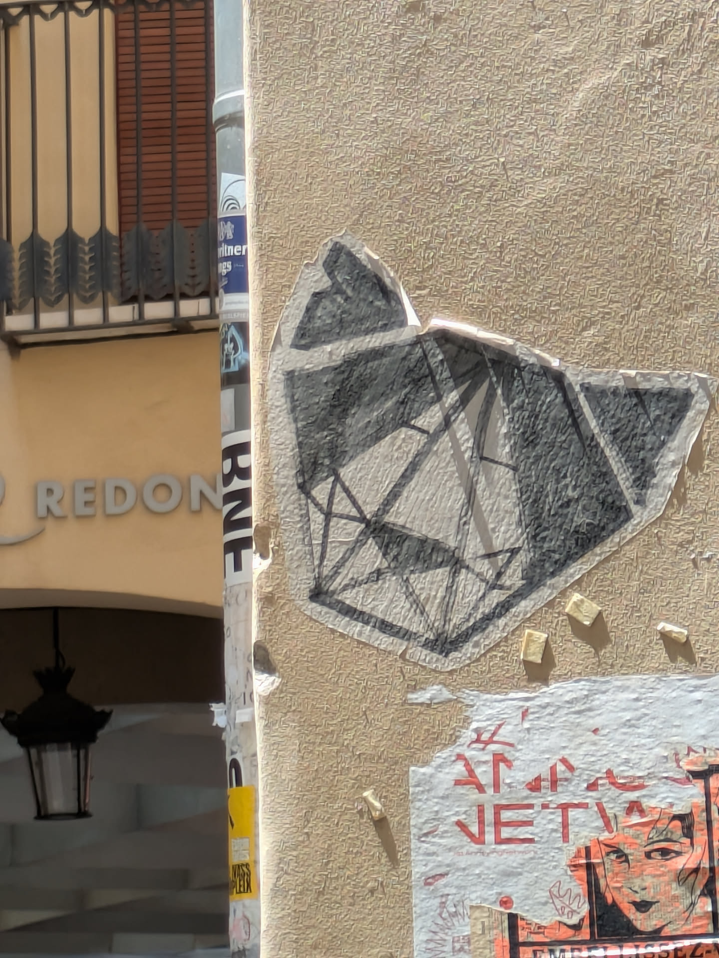 A geometric sticker adhered to a beige, textured wall. The sticker is grayscale and features a pattern of connected lines forming abstract shapes, resembling facets of a gem. Some small, rectangular pieces of paper or residue are scattered around the sticker. Below it, a partially torn poster with red text and graphic elements is visible.