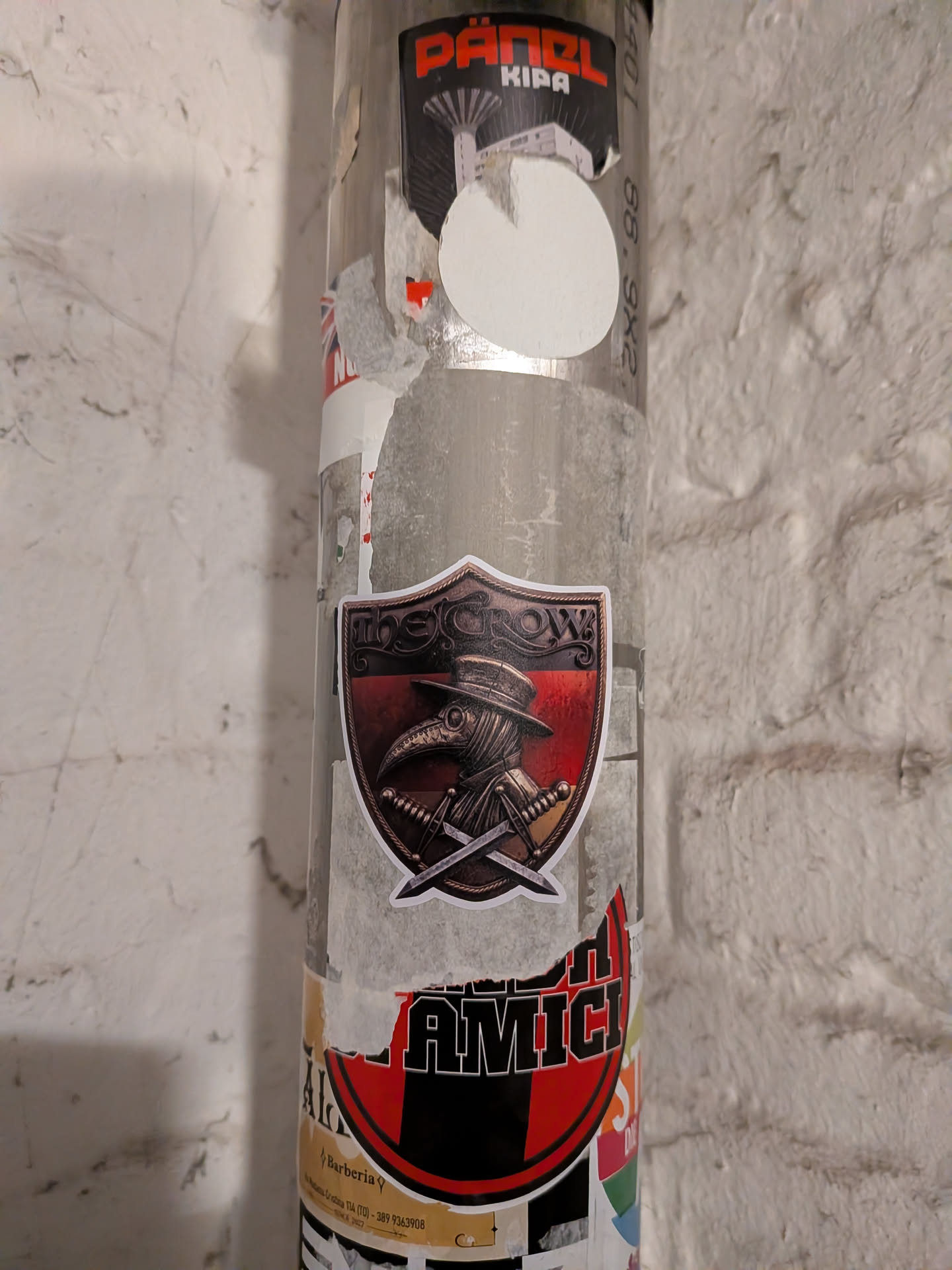 A die-cut, shield-shaped sticker featuring a detailed illustration with a bronze metallic, embossed aesthetic. At the top, an ornate header reads 'THE CROW'. Centered within the shield is a profile of a plague doctor wearing a wide-brimmed hat, beaked mask, and cowl, backed by dark red and black horizontal bands. In front of the figure are two crossed daggers. The sticker has a distinct white outline.