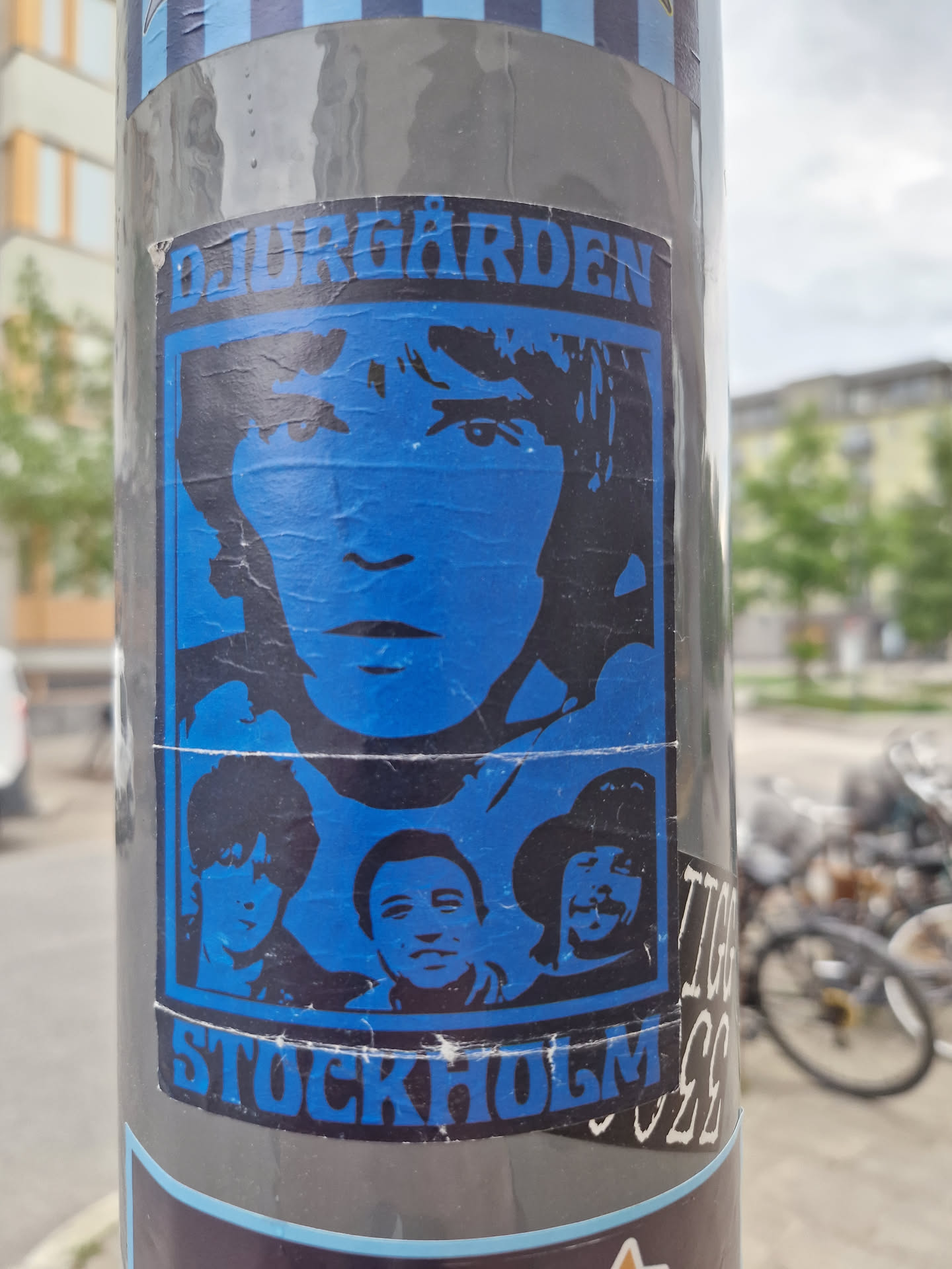 A sticker featuring a stylized blue portrait of what appears to be a young man, possibly a musician, with the text "DJURGÅRDEN" at the top and "STOCKHOLM" at the bottom.  Below the main portrait are smaller images of what seems to be a band or group of people. The sticker has a slightly worn look, showing signs of being affixed outdoors.