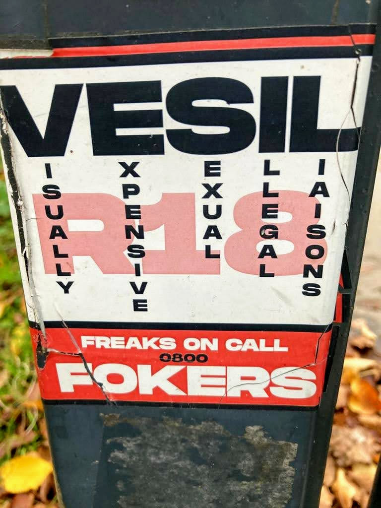 The sticker is a rectangular, aged advertisement affixed to a dark, possibly metal, pole. Its primary colors are white, black, and red, showing significant signs of wear with prominent creases, tears, and discoloration, particularly at the bottom and right edge. The top features a narrow red band above the large, bold, black text 'VESIL'. Below this, four vertical columns of black text spell out 'VISUALLY', 'EXPENSIVE', 'SEXUAL', and 'LIAISONS', forming an acronym related to 'VESIL'. Behind these words, a large, faded pinkish-red 'R18' is prominently displayed. The bottom section of the sticker consists of a wide red band containing white text: 'FREAKS ON CALL', a smaller '0800' number, and a very large, bold 'FOKERS' at the very bottom. The overall appearance is that of a crude, yet striking, adult-themed advertisement.
