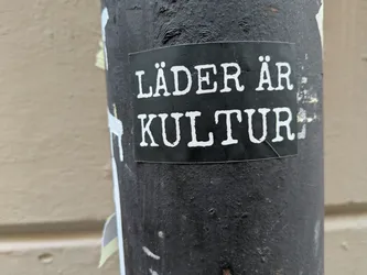 A black sticker with white text on it. The text says "Läder är kultur" which translates to "Leather is culture". The sticker is on a dark gray pole.