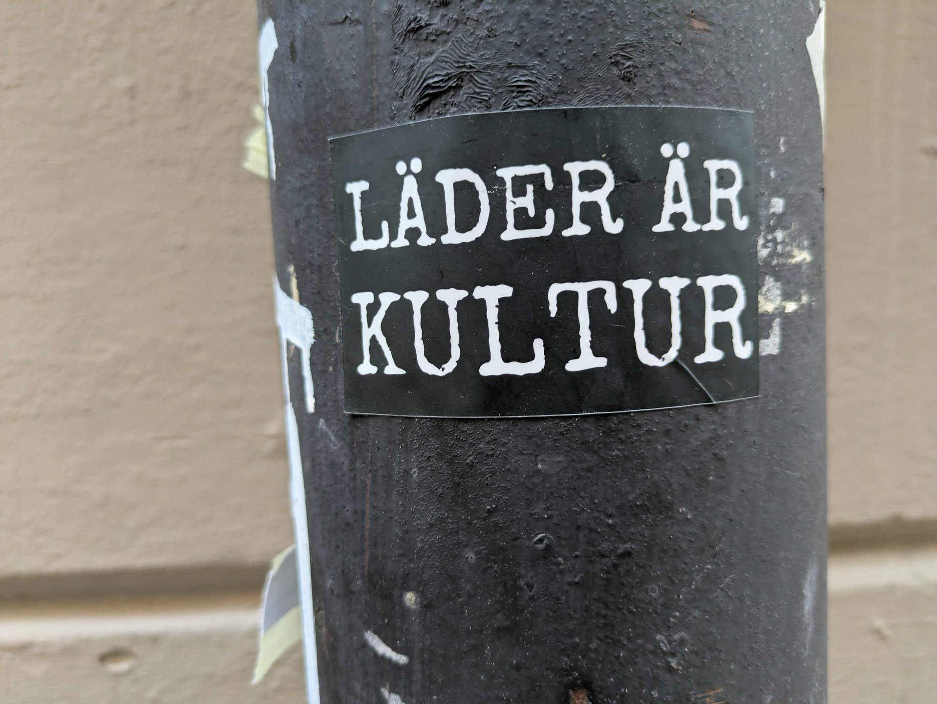A black sticker with white text on it. The text says "Läder är kultur" which translates to "Leather is culture". The sticker is on a dark gray pole.