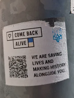 A white square sticker on a pole with the text "COME BACK ALIVE" framed in a black box with a heart on the left and the Ukrainian flag colors below. Next to it, the text says "WE ARE SAVING LIVES AND MAKING HISTORY ALONGSIDE YOU." It also contains a QR code and a stylized image.