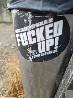 A round black and white sticker with the text "FUCKED UP!" in large letters.  The text "www.fuckedupberlin.de" is visible at the top, and #fuckedupberlin is at the bottom. An Instagram logo is also present. The sticker is affixed to a metal pole.