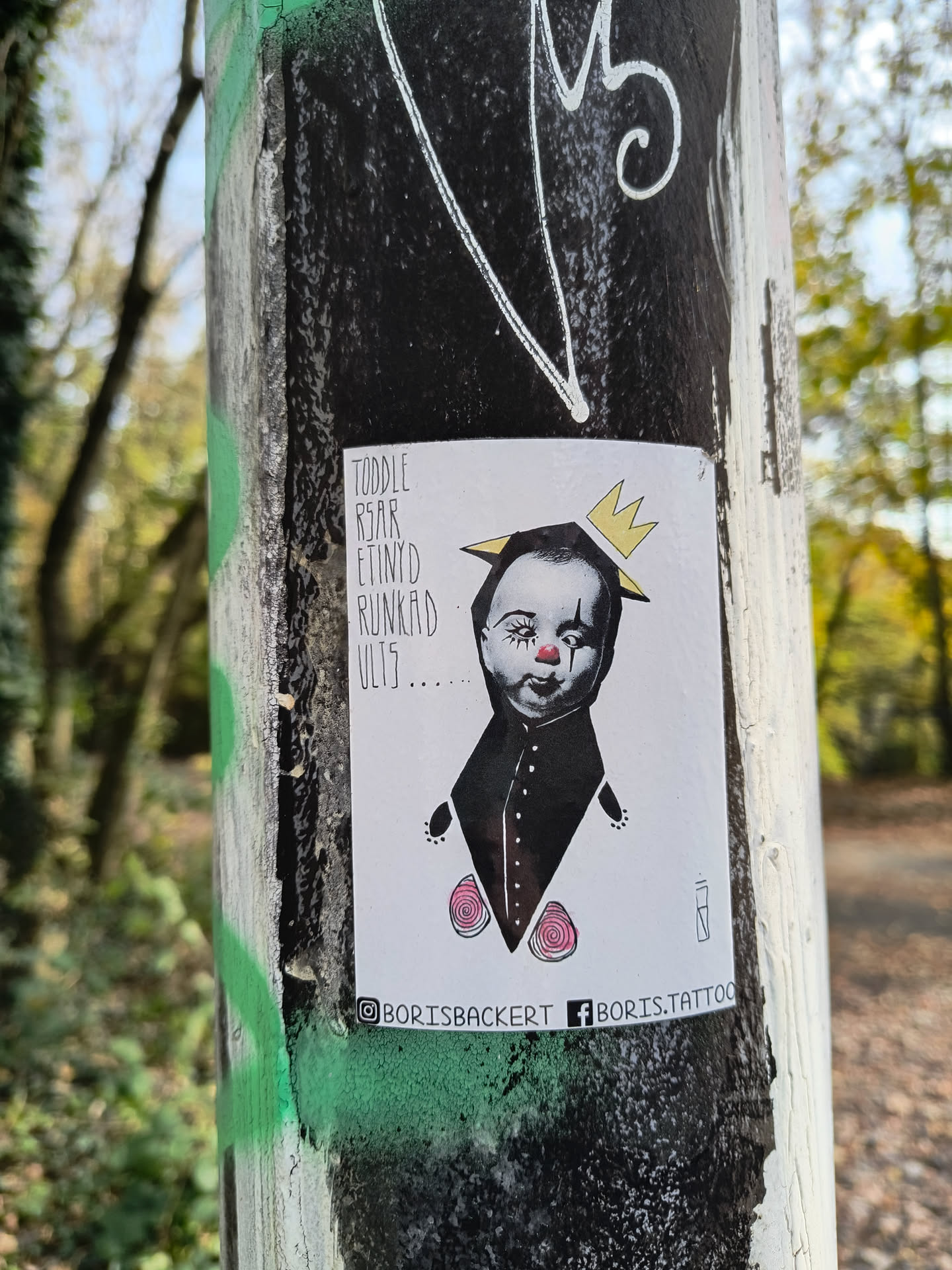 A sticker featuring an illustration of a toddler wearing a crown and a dark collared garment. The style is dark and slightly whimsical, with small pink rose-like details on the garment.  Text on the sticker includes an unclear phrase and social media handles.