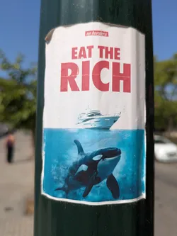 The sticker shows the text "Eat the Rich" above an image of a yacht sailing above the water, and an orca swimming below it. The sticker appears to be placed on a dark green pole.