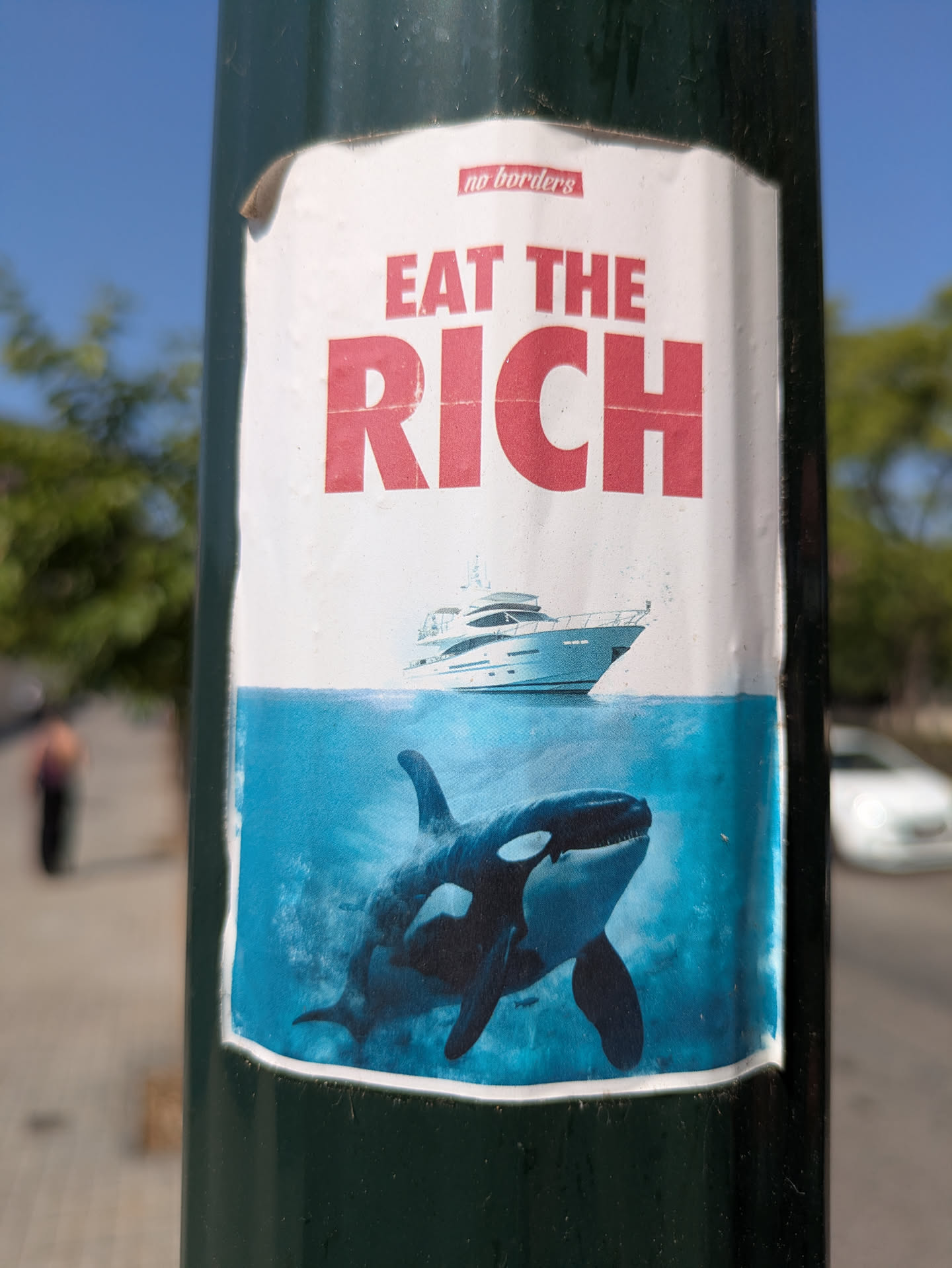 The sticker shows the text "Eat the Rich" above an image of a yacht sailing above the water, and an orca swimming below it. The sticker appears to be placed on a dark green pole.