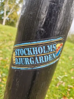 A worn sticker featuring the text "Stockholms Djurgården" in a vintage style. The sticker has a blue, yellow, and red color scheme and appears to be affixed to a dark-colored pole outdoors.