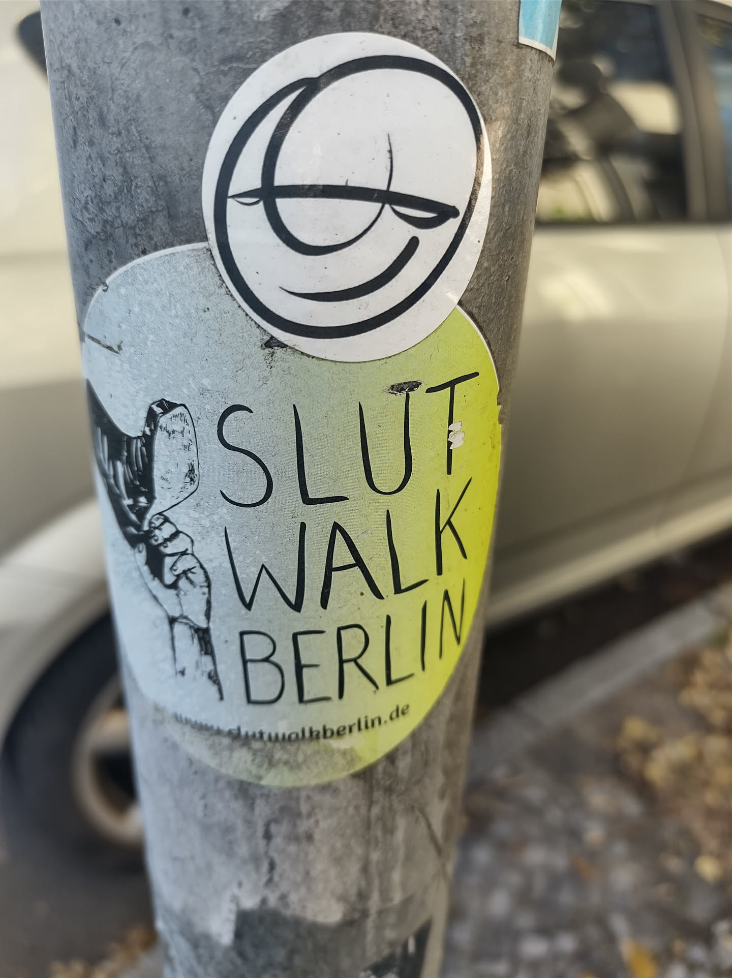 A sticker featuring the text "SlutWalk Berlin" and a small illustration of a hand.  There's also a separate smaller round sticker with a simple line drawing of a face above it.