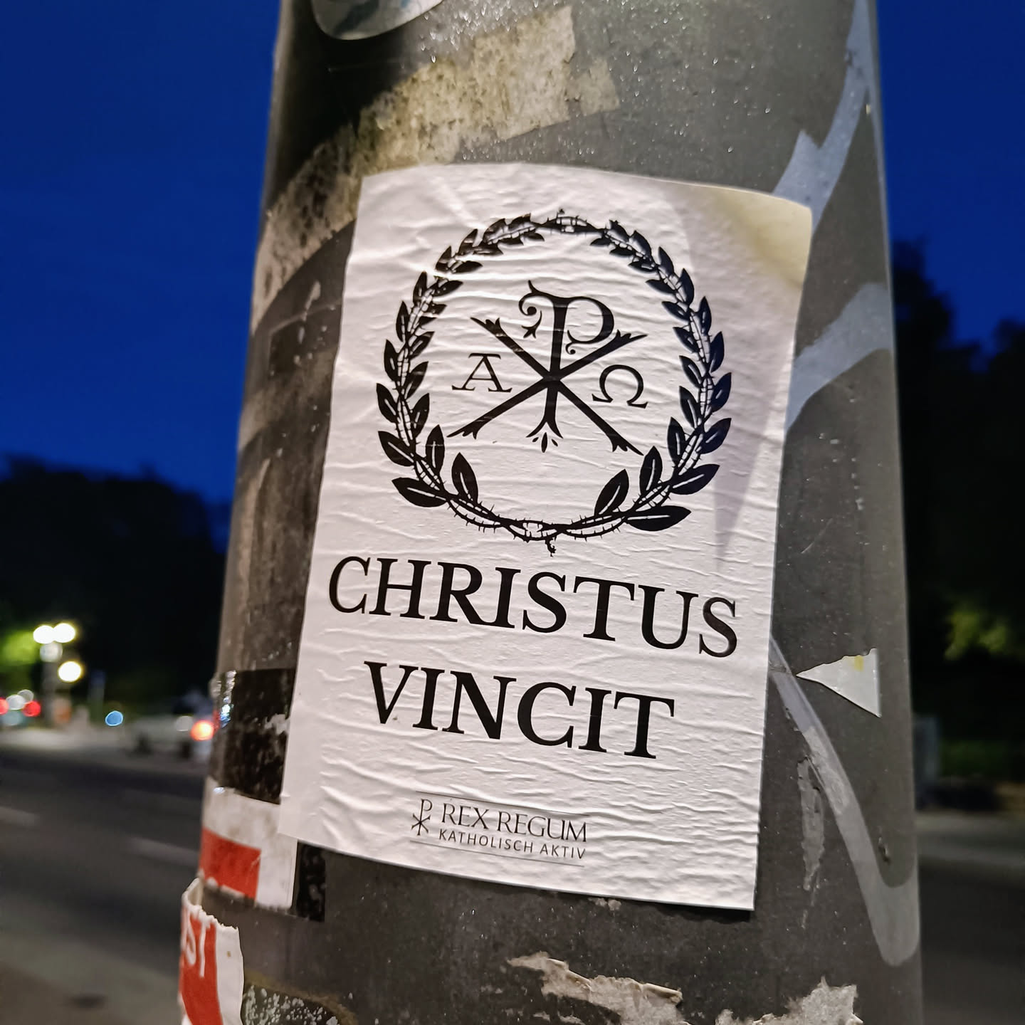 A sticker featuring the Chi Rho symbol within a laurel wreath, with the text "CHRISTUS VINCIT" and small print indicating "Rex Regum" and "Katholisch Aktiv".