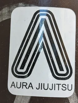 A white sticker with a black stylized letter 'A' and the text 'Aura Jiu Jitsu' underneath. The design is minimalist and geometric.