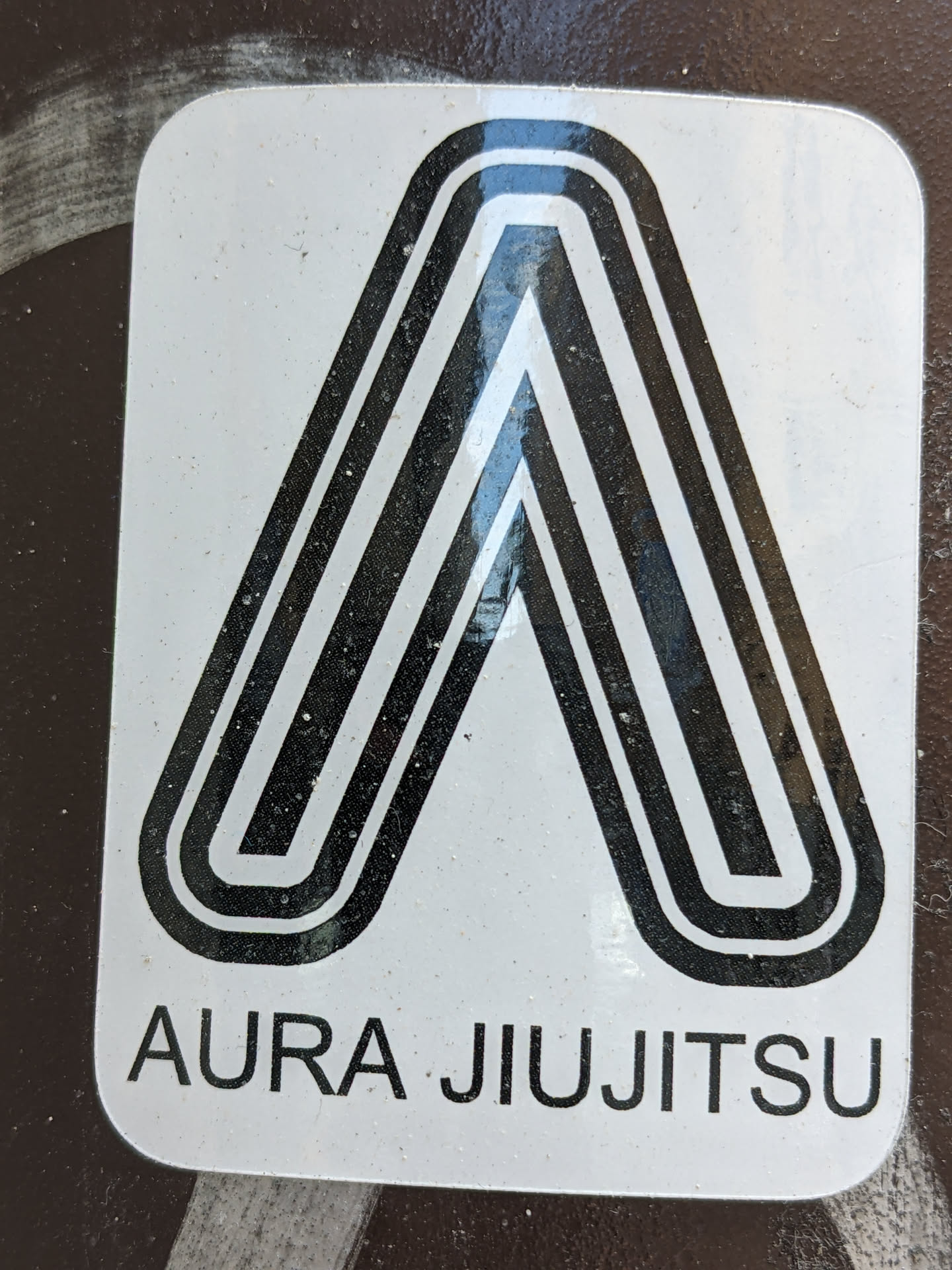 A white sticker with a black stylized letter 'A' and the text 'Aura Jiu Jitsu' underneath. The design is minimalist and geometric.