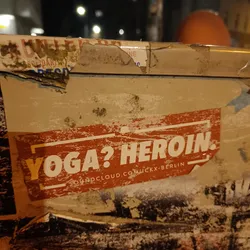 A worn sticker on a wall with the text "Yoga? Heroin." and a SoundCloud link. The sticker is orange with white text.