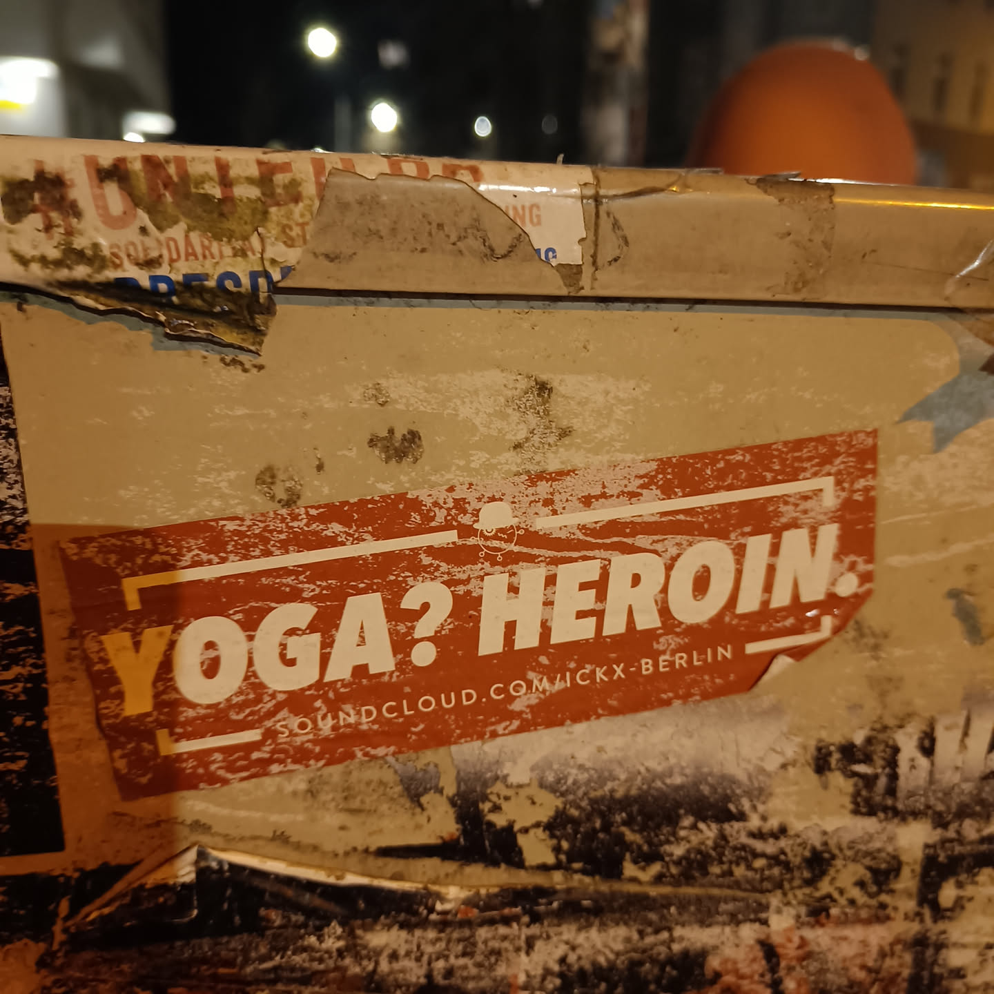 A worn sticker on a wall with the text "Yoga? Heroin." and a SoundCloud link. The sticker is orange with white text.