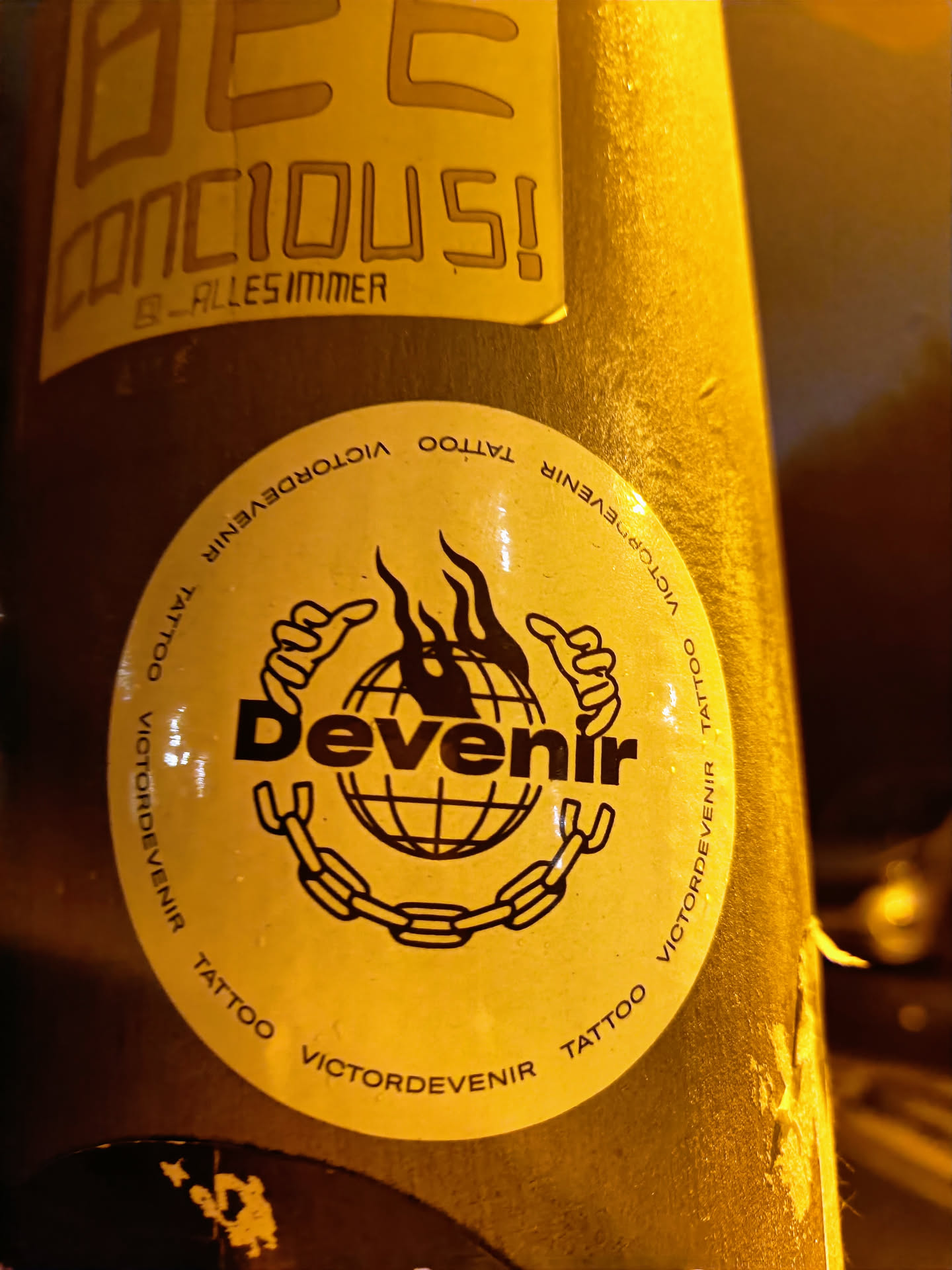 Round sticker with the word 'Devenir' in bold text inside a circular emblem featuring a globe encircled by a chain, all set against a yellow background. The text 'VictorDevenir Tattoo' is repeated around the outer edge of the circle.  The style is bold and graphic.