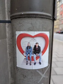 Street sticker A rectangular photographic sticker attached to a metal post, showing a young woman in a blue jacket and beanie and a young man in a black jacket and jeans. They are seated side by side on a white bench enclosed within a large, bulbous red heart-shaped frame. Faint decorative scrollwork and the text 'TEMPORAR' appear in the background behind them.