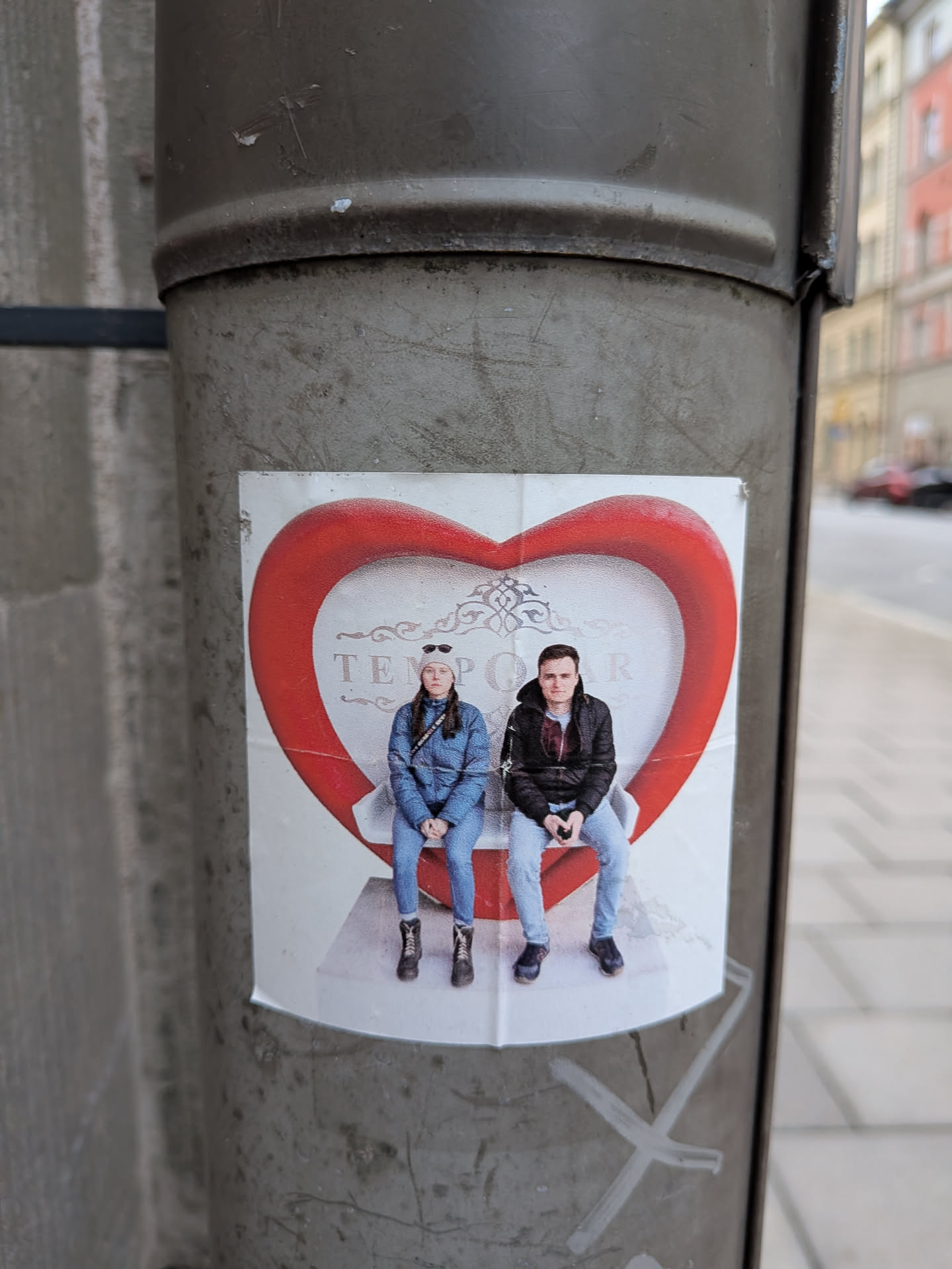 A rectangular photographic sticker attached to a metal post, showing a young woman in a blue jacket and beanie and a young man in a black jacket and jeans. They are seated side by side on a white bench enclosed within a large, bulbous red heart-shaped frame. Faint decorative scrollwork and the text 'TEMPORAR' appear in the background behind them.
