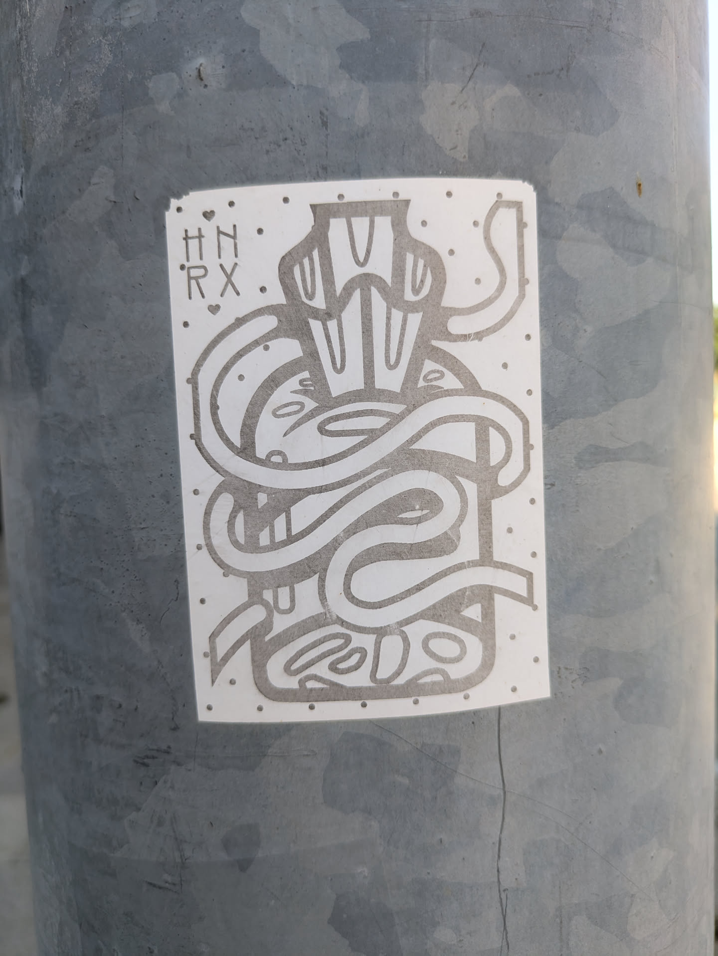 A monochrome sticker adhered to a gray pole features an abstract design. The drawing includes what appears to be a container with swirling elements and organic shapes within it. The letters 'HN' above 'RX', separated by small hearts, are located in the upper-left corner of the sticker.