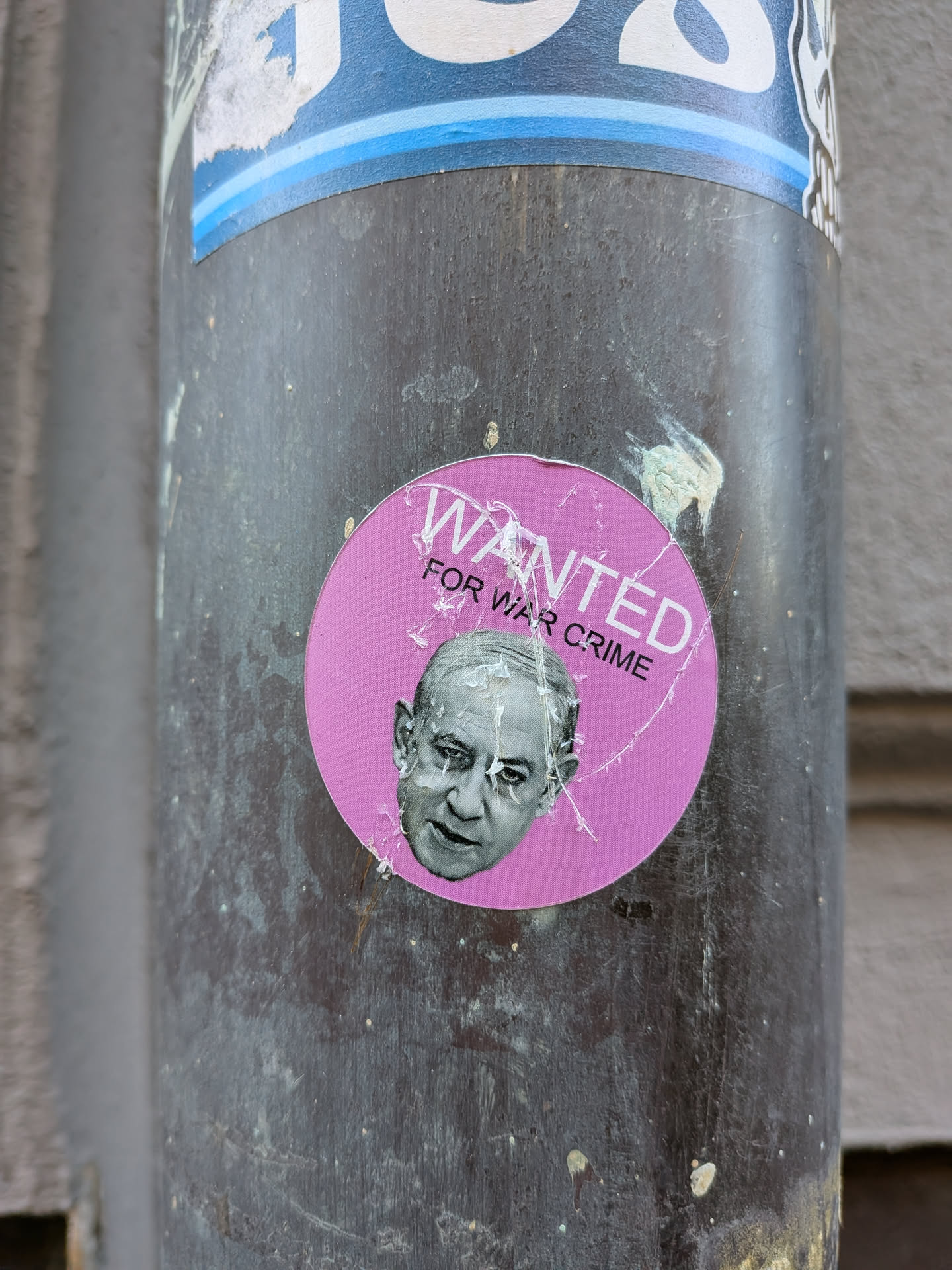 The sticker is circular with a vibrant pink background. In the upper portion, the word "WANTED" is prominently displayed in large, white, sans-serif capital letters with a slight curve. Below it, in smaller white capital letters, is the text "FOR WAR CRIME". Centered below the text is a grayscale portrait of an elderly man with a stern, displeased expression. The sticker shows signs of wear and tear, with visible creases and peeling edges, indicating it has been affixed to the surface for some time. Some white and light blue elements from another sticker are visible above it.