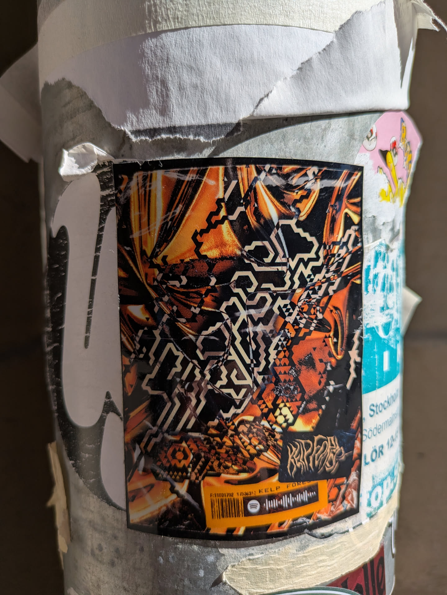 A close-up shot shows an abstract sticker with a geometric pattern of orange and black. The bottom of the sticker shows a Spotify code and the text reads "KELP FOREST". Another text on the sticker says "RIPFACE". The sticker is stuck on a pole and appears to be part of a collection of other stickers.
