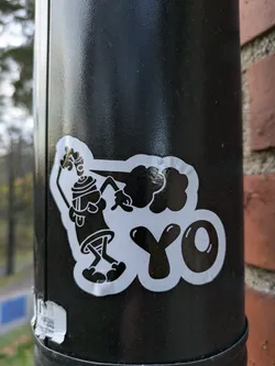 A close-up shot of a sticker on a black pole. The sticker features a cartoon character holding a spray can and the word "YO" in bold letters. The style is reminiscent of classic graffiti art.