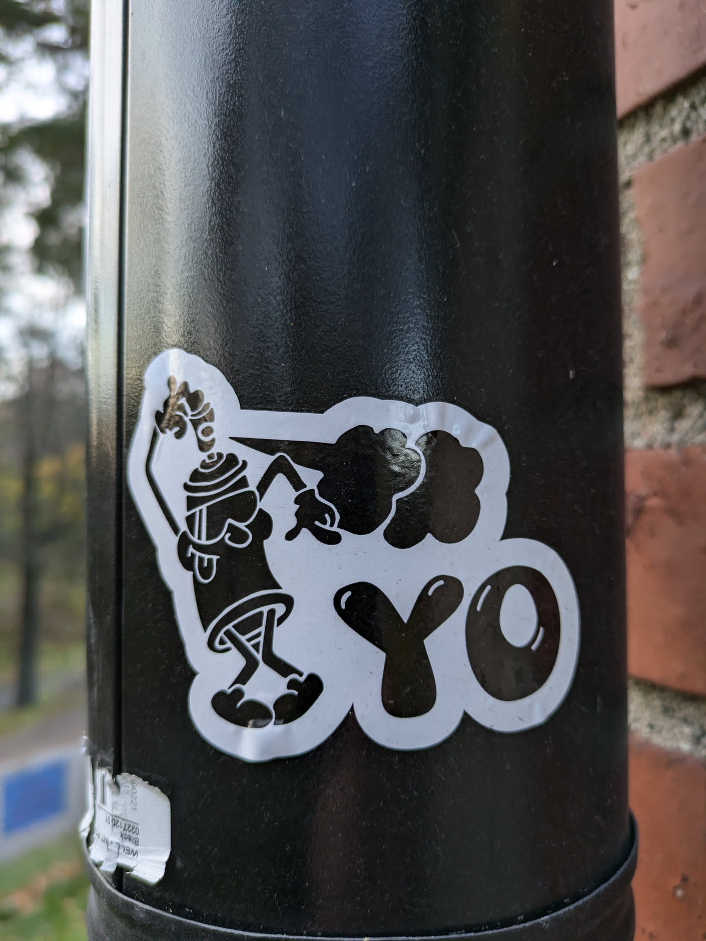 A close-up shot of a sticker on a black pole. The sticker features a cartoon character holding a spray can and the word "YO" in bold letters. The style is reminiscent of classic graffiti art.