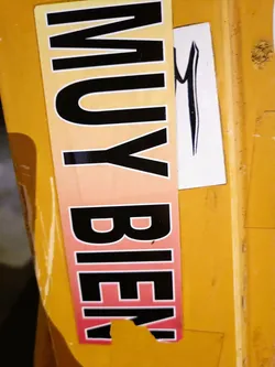 A sticker with the words "MUY BIEN" in black letters on a gradient orange-pink background.  The sticker is slightly damaged at the bottom. There is a smaller, separate sticker with a stylized black 'M' next to it.